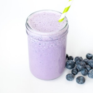 A clear glass bowl of blueberry milk against a white background with blueberries in front and a green striped straw coming out of the glass.
