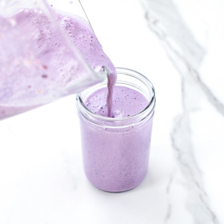 Clear glass blender pouring blueberry milk into a clear glass jar against a marble countertop.