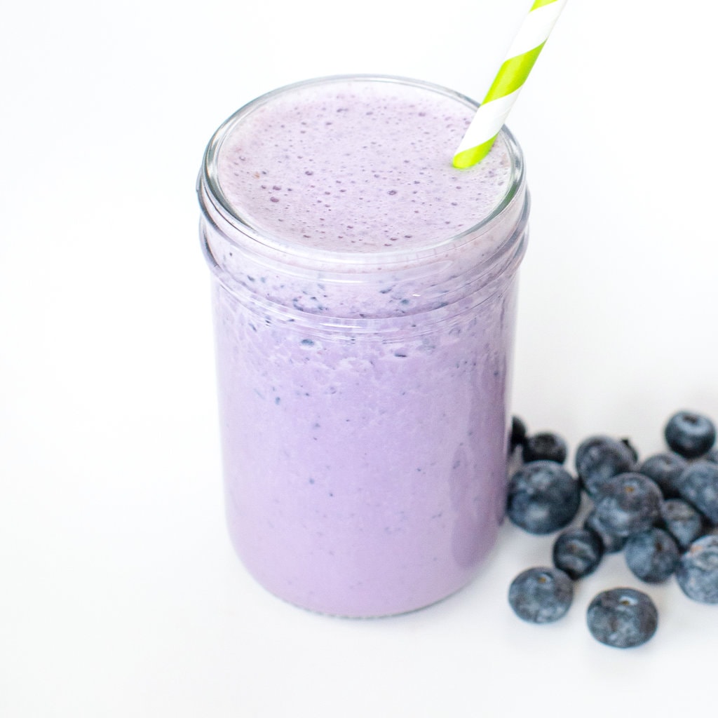 A clear glass bowl of blueberry milk against a white background with blueberries in front and a green striped straw coming out of the glass.