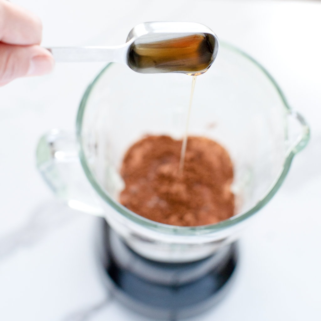 And pouring in Maple syrup into a clear blender on a white marble countertop.