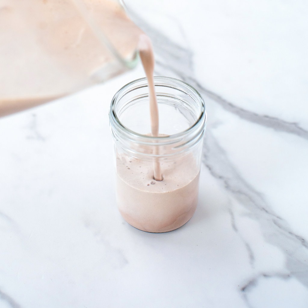 Clear blender pouring chocolate milk into a clear glass on a marble counte