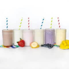 A line of clear glass cups filled with different flavors of milk – chocolate milk, strawberry milk, vanilla milk, mango milk, blueberry milk, and mango milk with colorful straws coming out against a white background.
