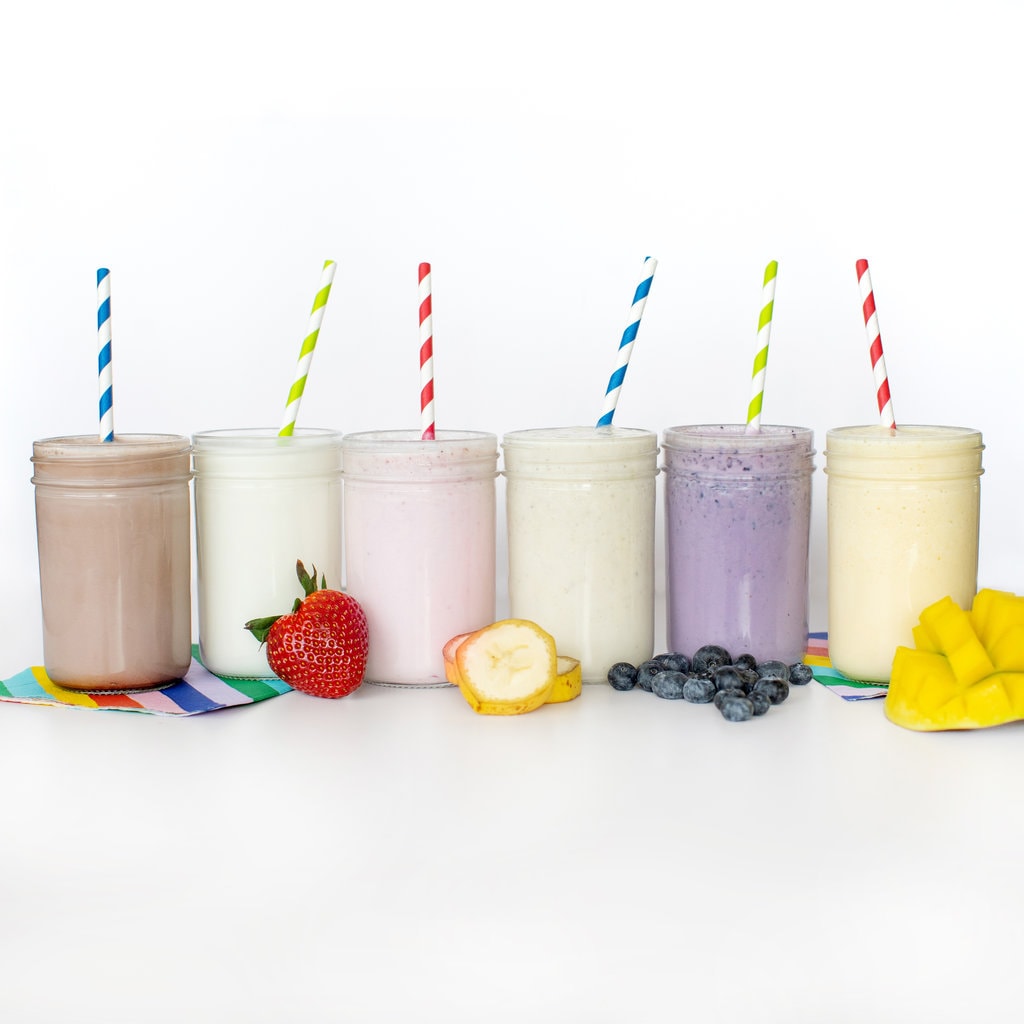 A line of clear glass cups filled with different flavors of milk – chocolate milk, strawberry milk, vanilla milk, mango milk, blueberry milk, and mango milk with colorful straws coming out against a white background.