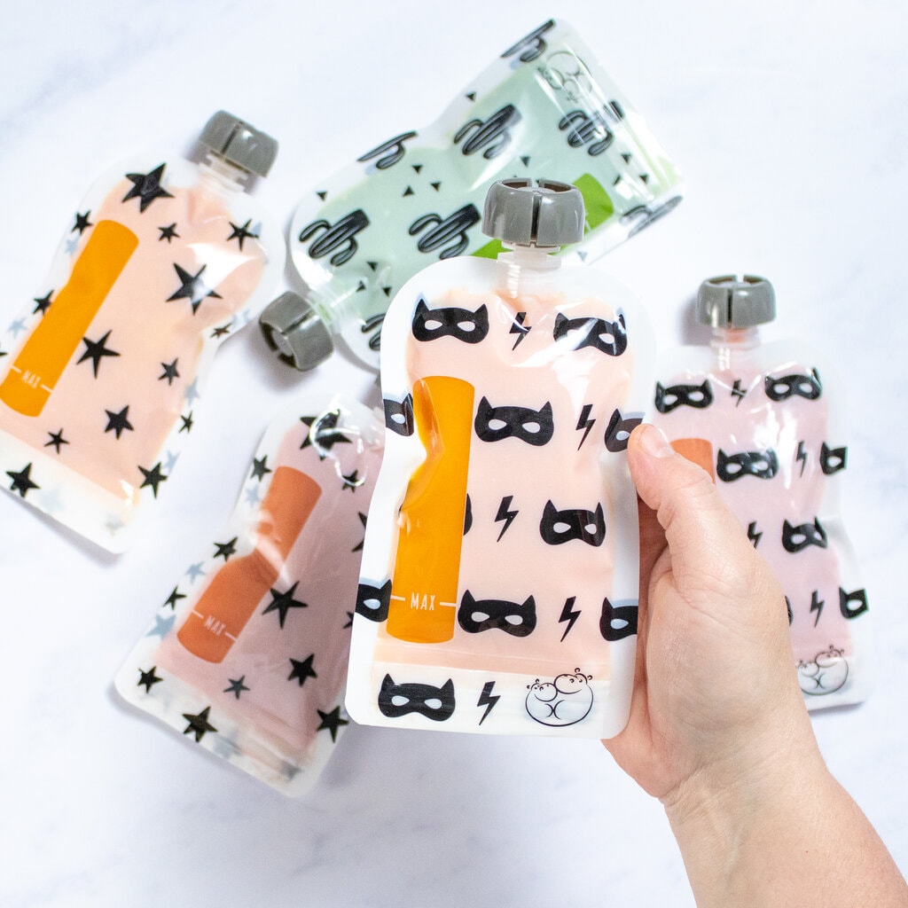 A hand holding a baby food pouch with an orange sweet potato purée with a variety of baby food pouches on a marble countertop.