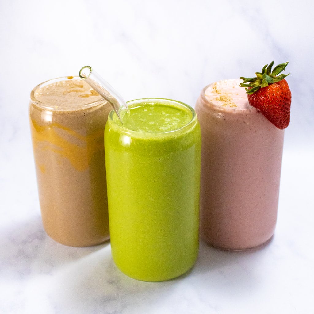 Three clear glasses full of smoothies for breast-feeding moms. One is green spinach and peach, one is pink which is a strawberry yogurt, and the other is chocolate peanut butter. All of his movies are against a white marble background.