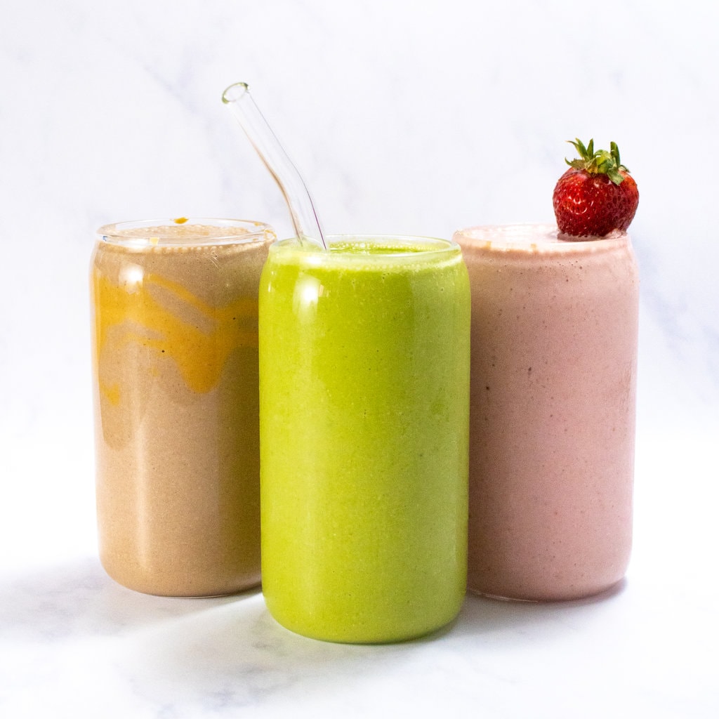 Three clear glasses full of smoothies for breast-feeding moms. One is green spinach and peach, one is pink which is a strawberry yogurt, and the other is chocolate peanut butter. All of his movies are against a white marble background.