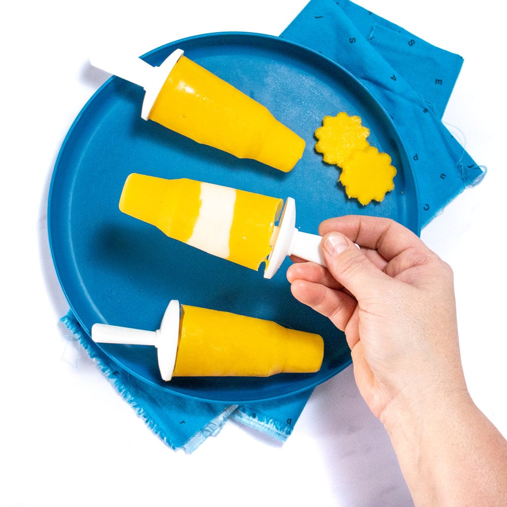 A hand holding a mango popsicle against a dark blue plate with more mango popsicles in a blue napkin.