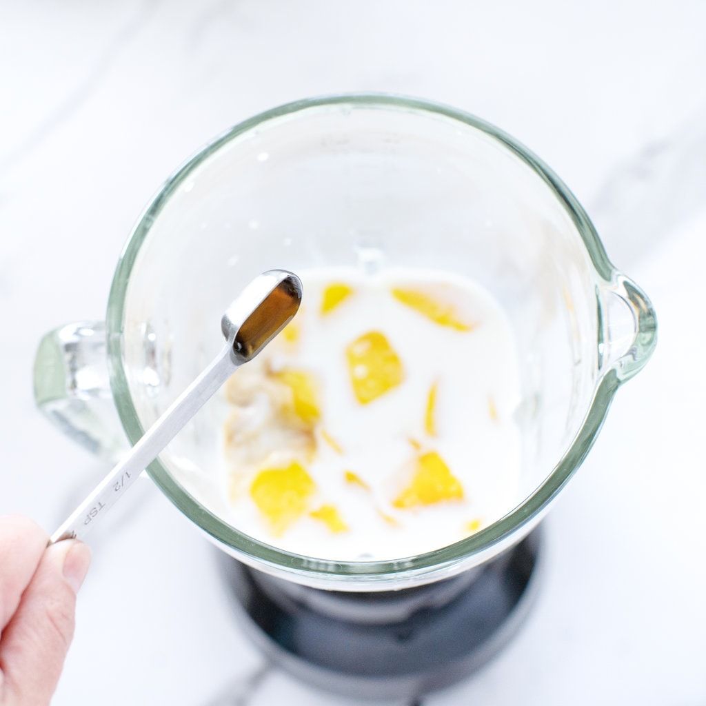 A clear glass blender with mango, milk and maple syrup with a hand pouring in vanilla extract.
