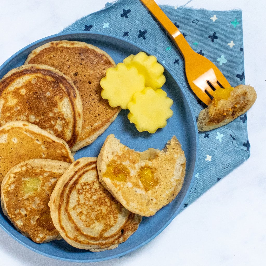 And a close look at a blue kids plate full of mango pancakes with mango shapes cut out an orange fork, a blue colorful napkin and a white background.