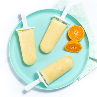 A teal kids plate with orange Creamsicle's and some orange slices on top of a white marble countertop.