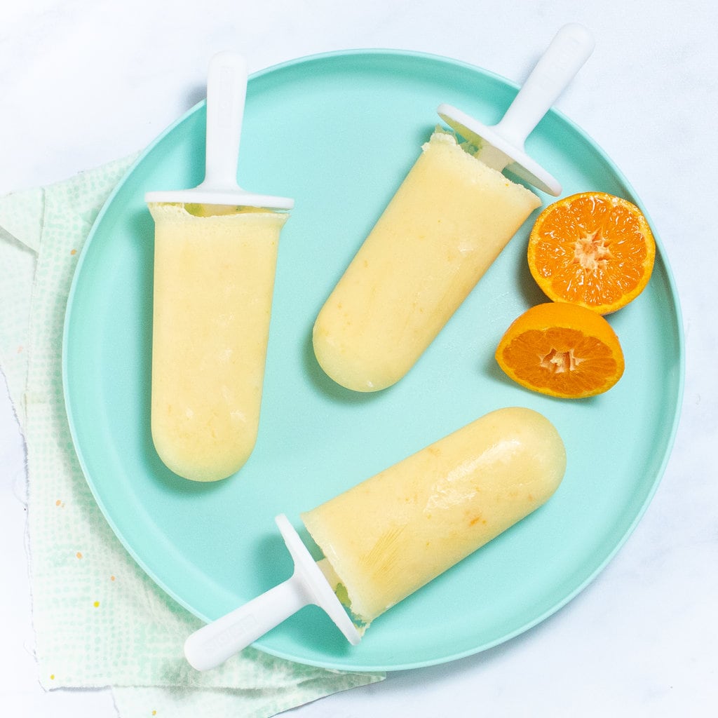 A teal kids plate with orange Creamsicle's and some orange slices on top of a white marble countertop.