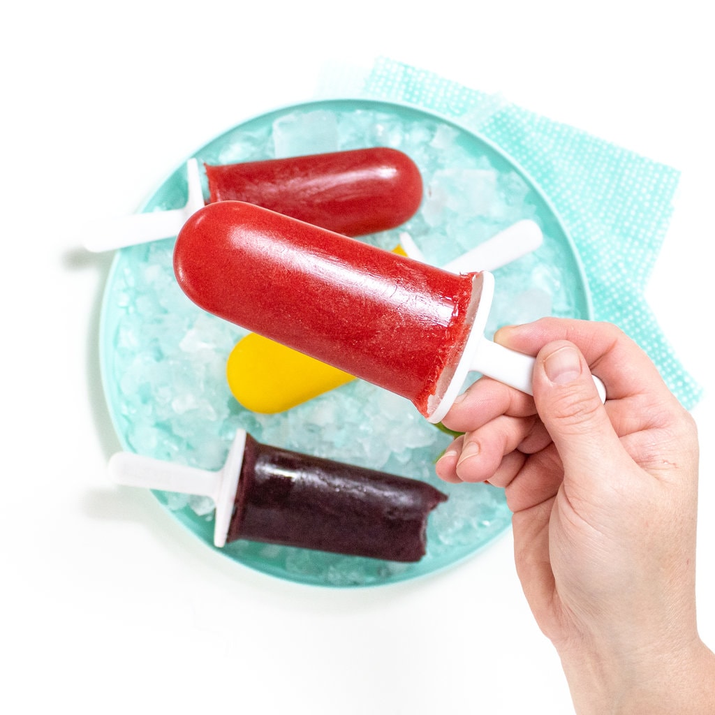 Hand holding a bright pink strawberry popsicle over a plate of different fruit popsicles.