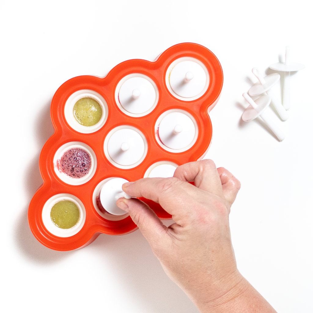 A hand reaching to put in a popsicle stick on a red and white popsicle mold with grape Popsicles inside.