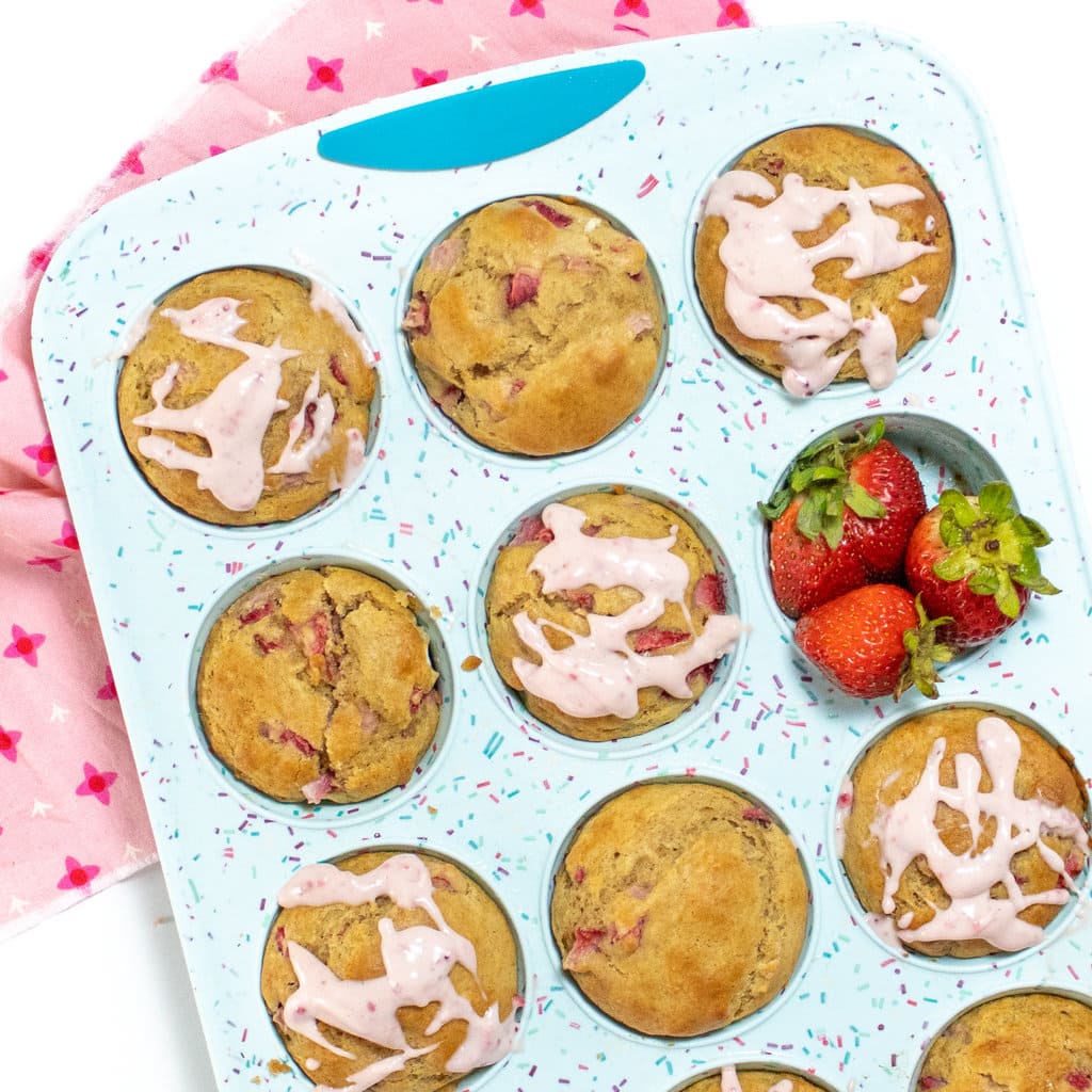 A blue muffin tin full of strawberry muffins with a cream cheese, strawberry, drizzle with a side of strawberries, a pink napkin, and a white background.