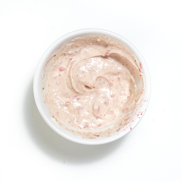 Small white bowl, full of cream cheese, strawberry icing on a white countertop.