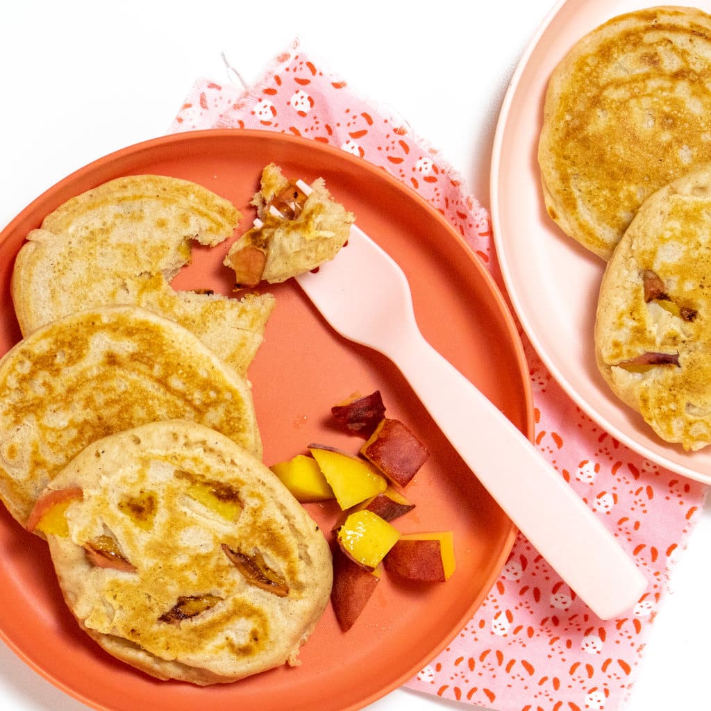 Two pink kids plates with peach pancakes and a pink fork with a pink napkin against a white background.