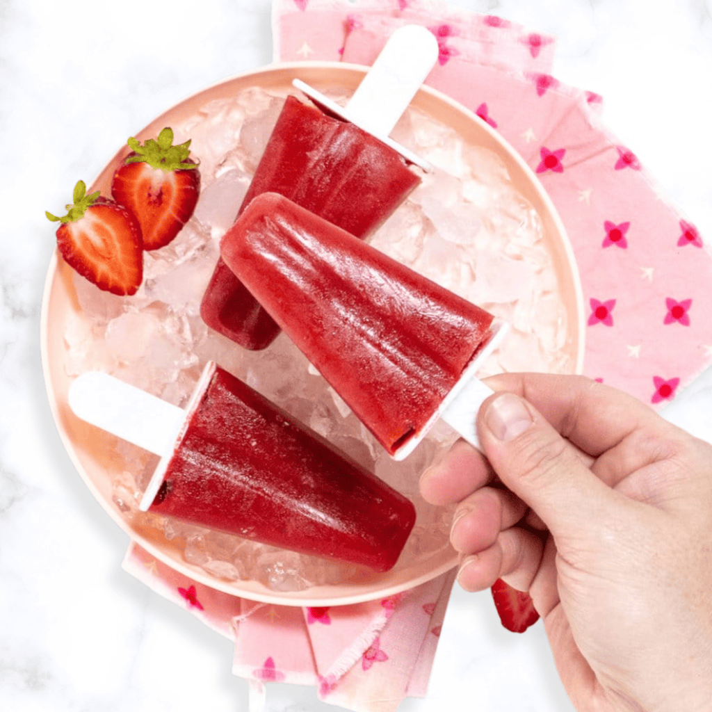 A pink plate full of ice chips and 3 strawberry popsicles with my hand holding one over the plate. The plate is sitting on my white marble kitchen counter. 