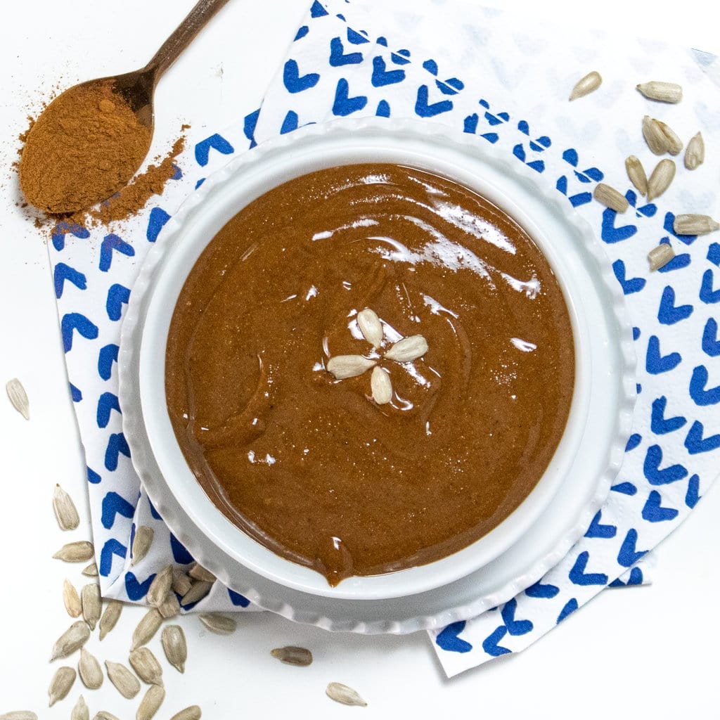 A bowl full of sunflower butter, on a white countertop, with a white and blue napkin with sunflower seeds scattered about.