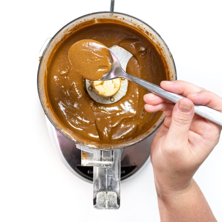 A hand, holding up a spoonful of smooth, sunflower seed, butter, over a food processor against a white background.