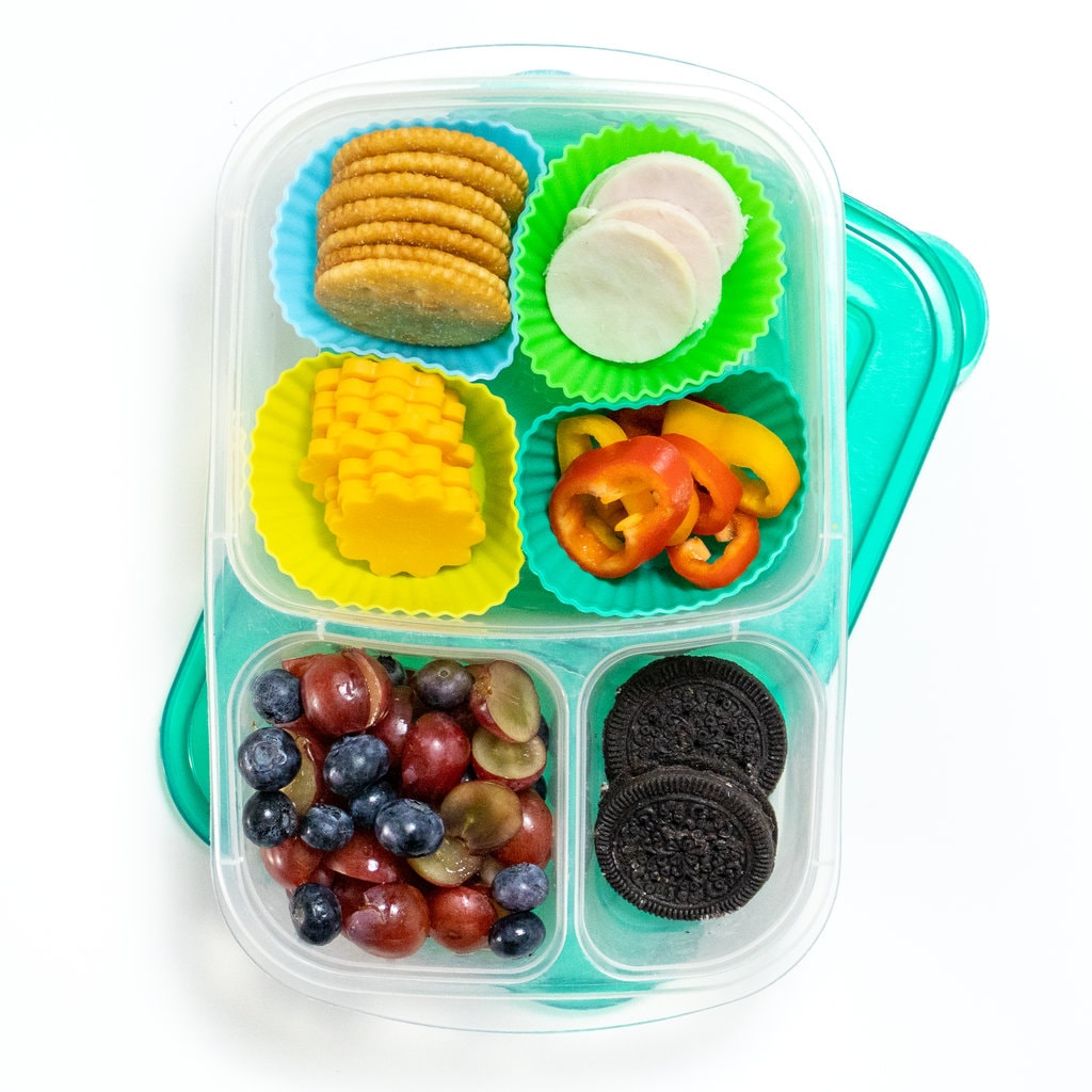Colorful kids lunch box with lots of different sections that are filled in with turkey, cheese, crackers, veggies and fruits.