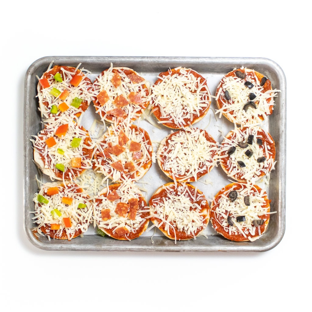 Mini bagels with sauce, cheese and different toppings on a baking sheet on a white counter.