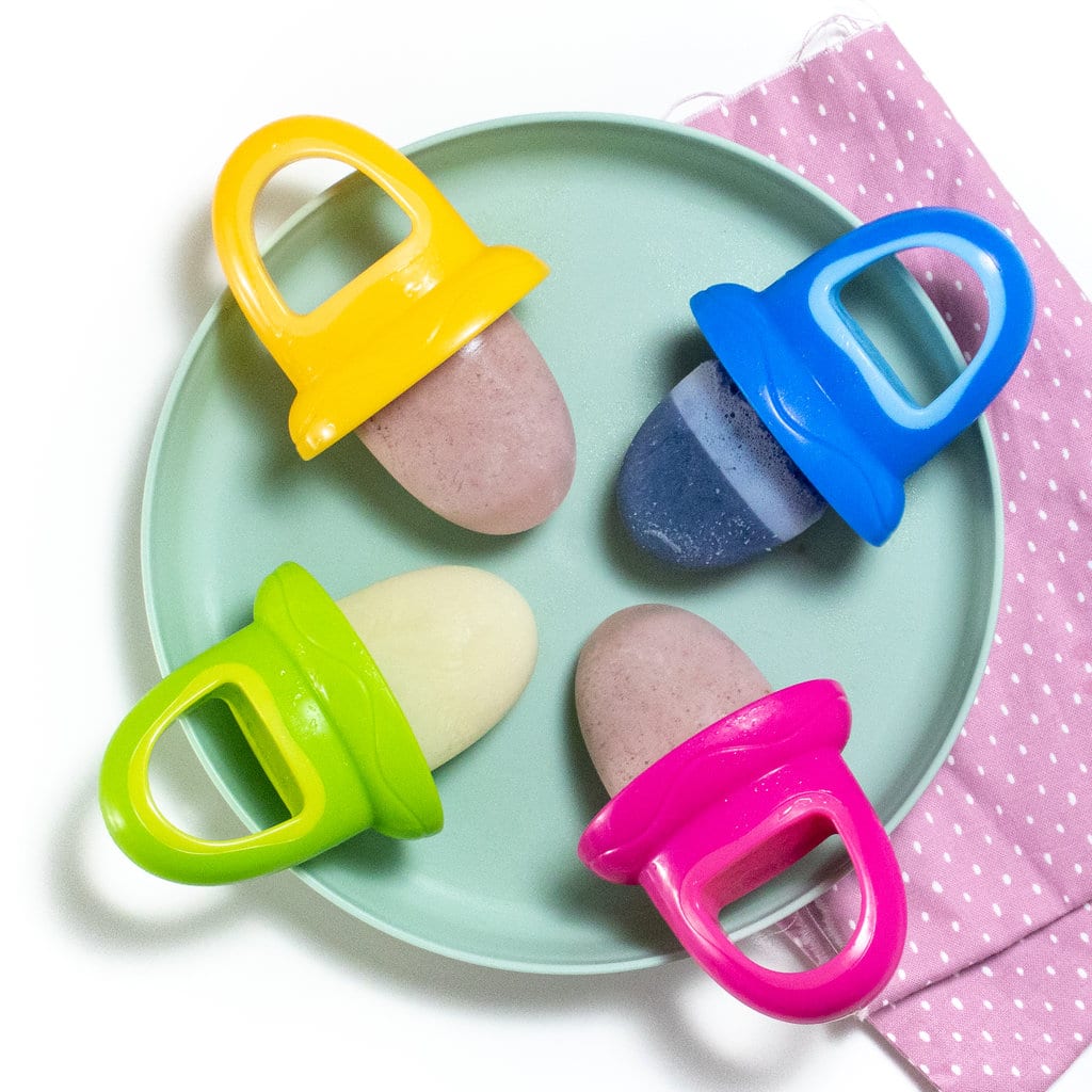 Teal baby plate with different flavors of breast milk popsicles on colorful popsicle sticks sitting on a purple napkin on a white counter. 