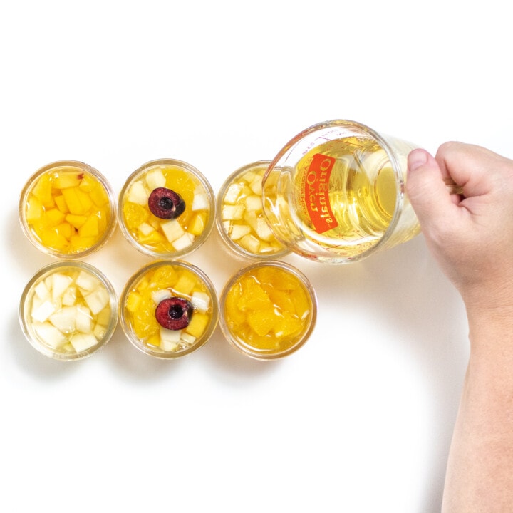 Small jars filled with an assortment of fruit with a hand pouring in juice to the top.