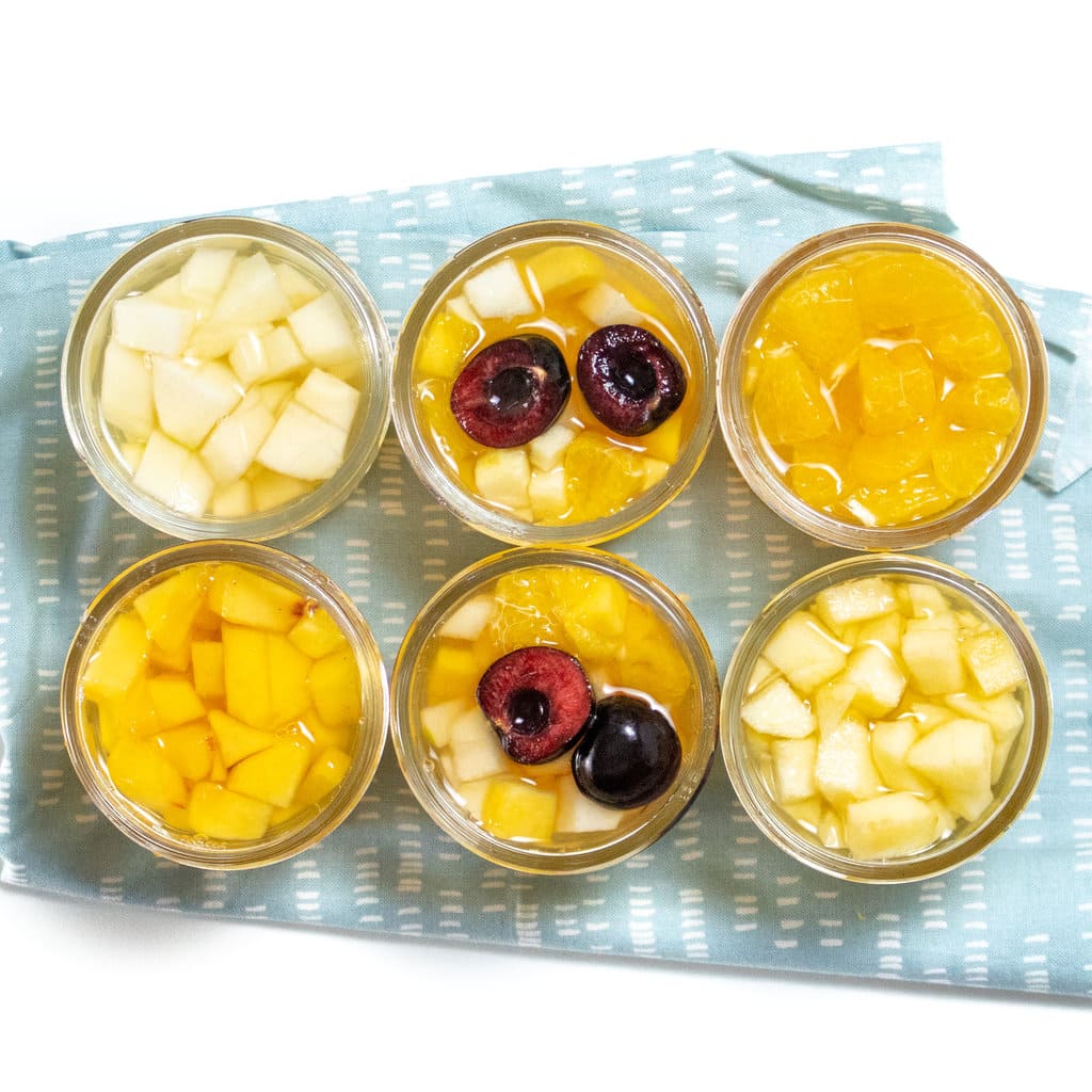6 small jars full of different fruit on top of a blue napkin sitting on a white counter.
