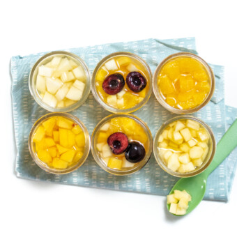 6 jars of fruit on a blue napkin with a green spoon resting next to one of them.