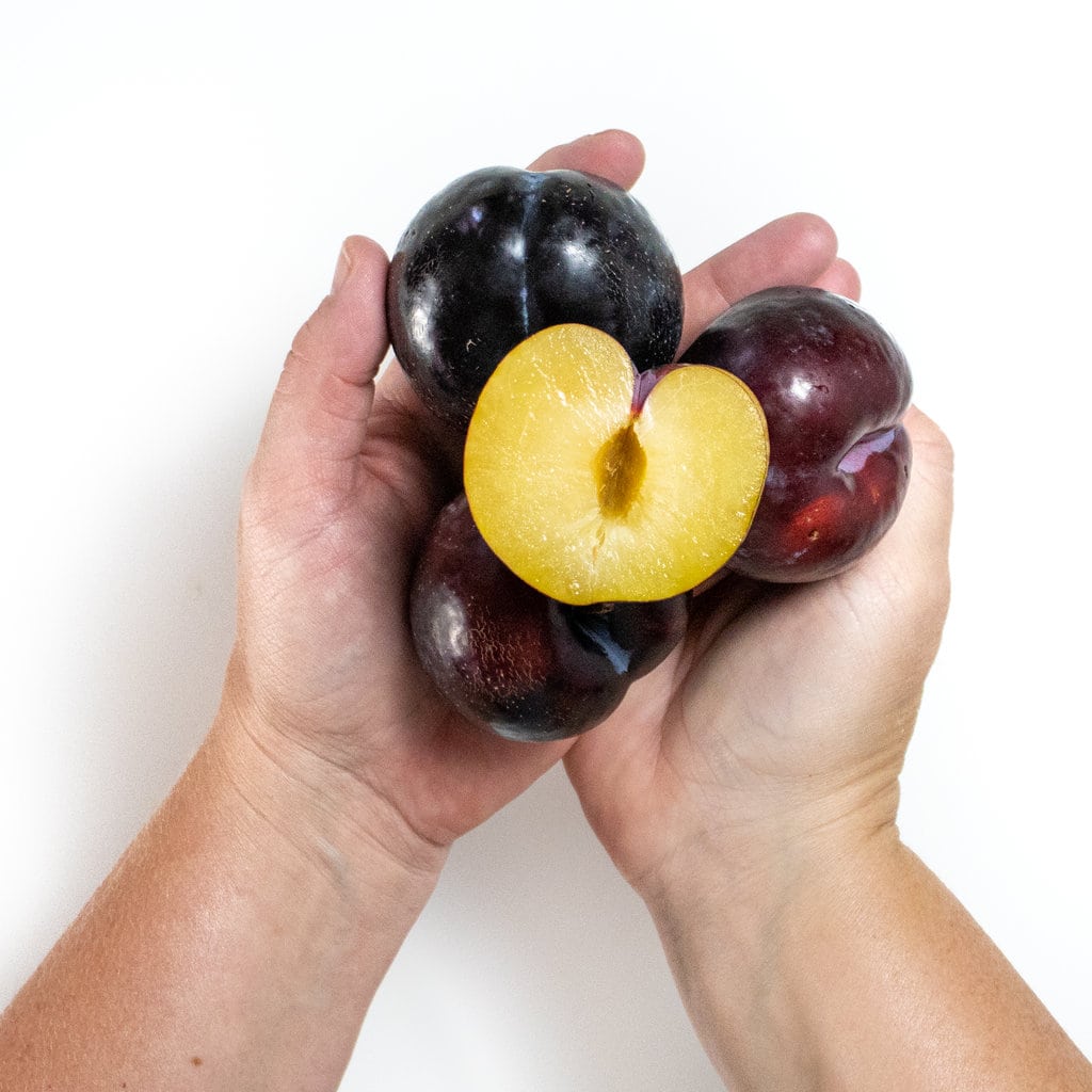Two hands holding a bunch of plums, the top one cut in half with the pit removed.