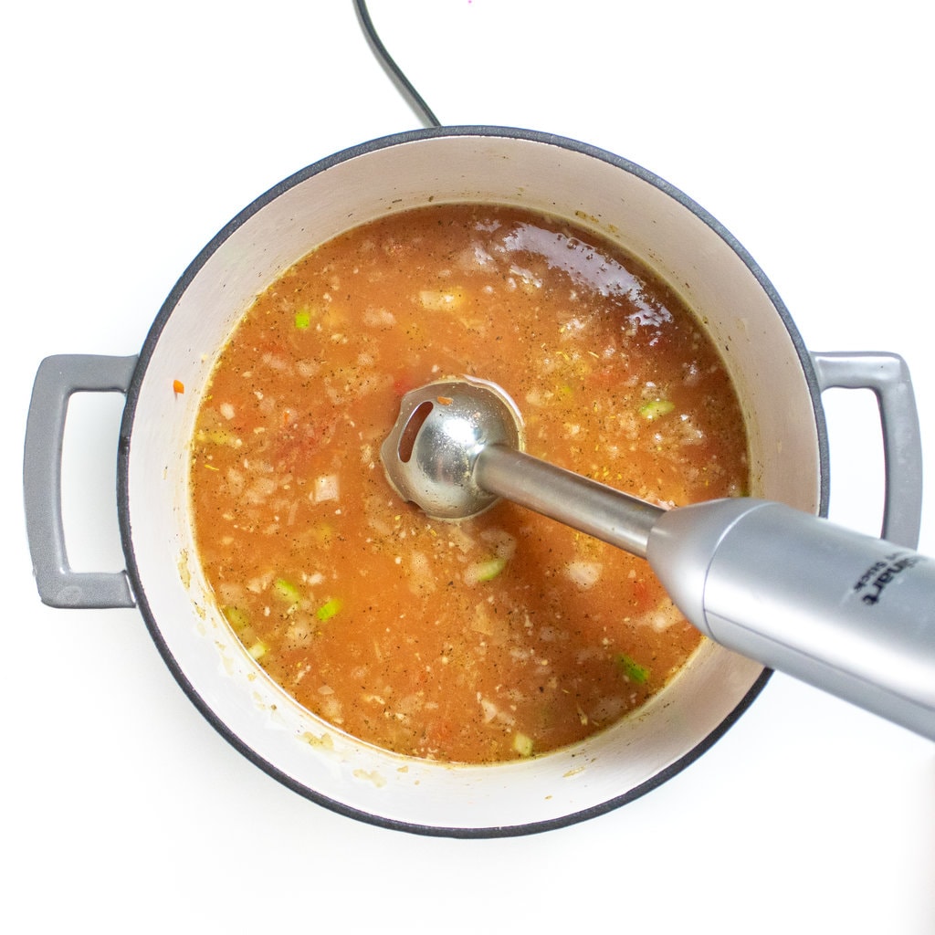 A large gray sock pot on the white counter with ingredients for alphabet pasta with an immersion blender ready to blend them up so the veggies are hidden.