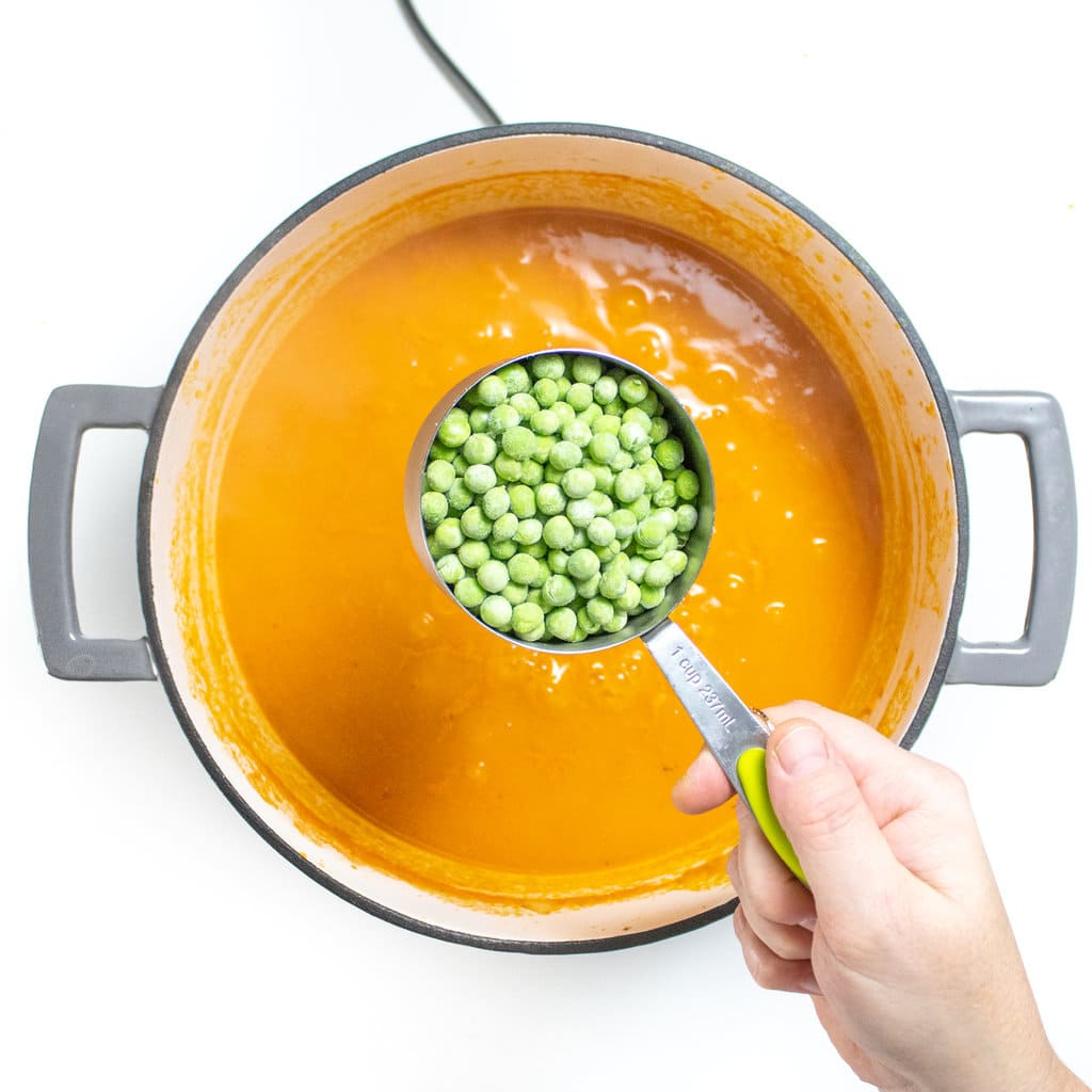 A large gray stock pot on a white counter, full of veggie broth and alphabet pasta with a hand holding a cup of frozen pe