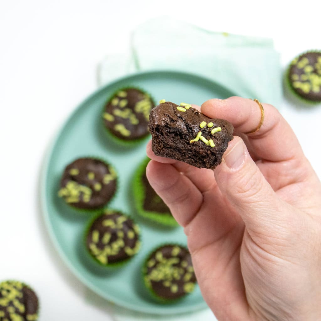A hand holding up a brownie bite that has been in and a half over a plate, full of brownie bites, and a tail napkin.