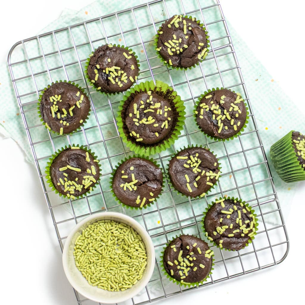 Hey, small, silver clean, rag with mini avocado, brownie bites, and a small white bowl, full of green sprinkles with a teal and blue napkin over a white counter.