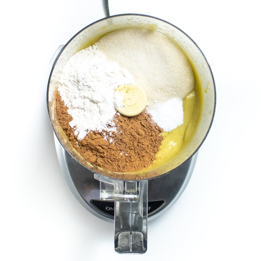 Food processor on the white counter with ingredients for avocado brownies.