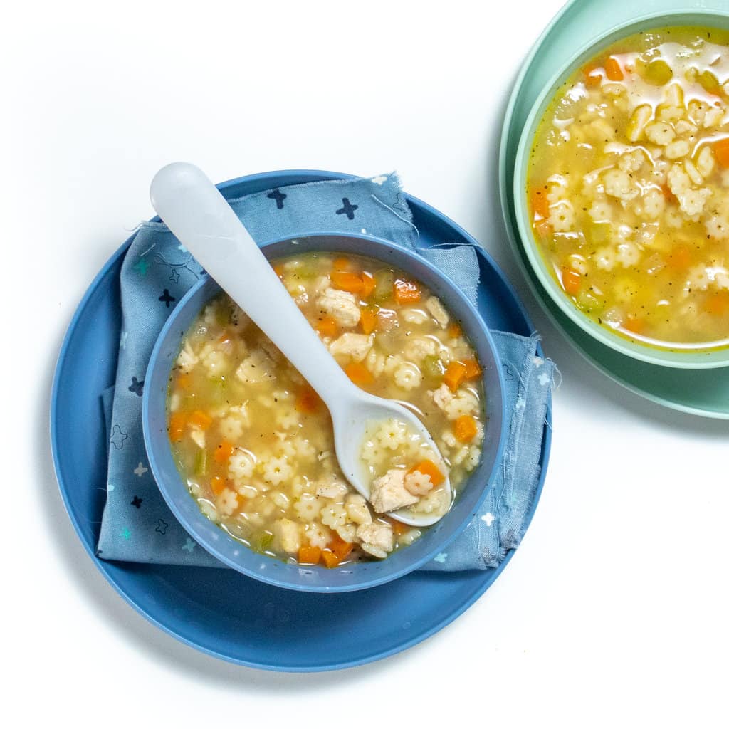Two sets of blue and teal kids, plates and bowls with a colorful napkin. Each of the bowls is filled with chicken and star soup. The blue bowl has a blue spoon, icing on top with star, pasta, diced chicken and carrots inside.