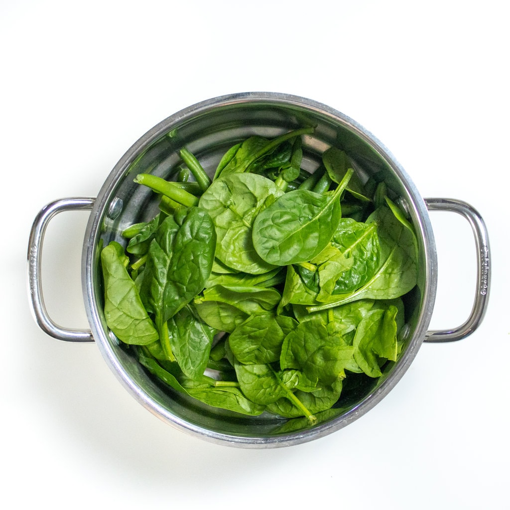 Silver steamer basket on a white countertop with fresh baby spinach, and green beans.