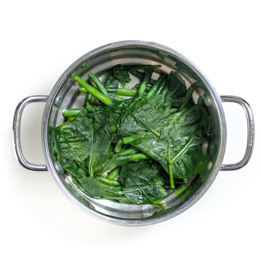 Silver steamer basket on the white countertop with steamed spinach and green beans.