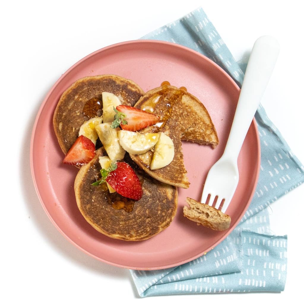 Pink kids played on a white countertop with three whole wheat pancakes, topped with chopped strawberries and bananas it with a drizzle of maple syrup with a white fork cut into the pancake.