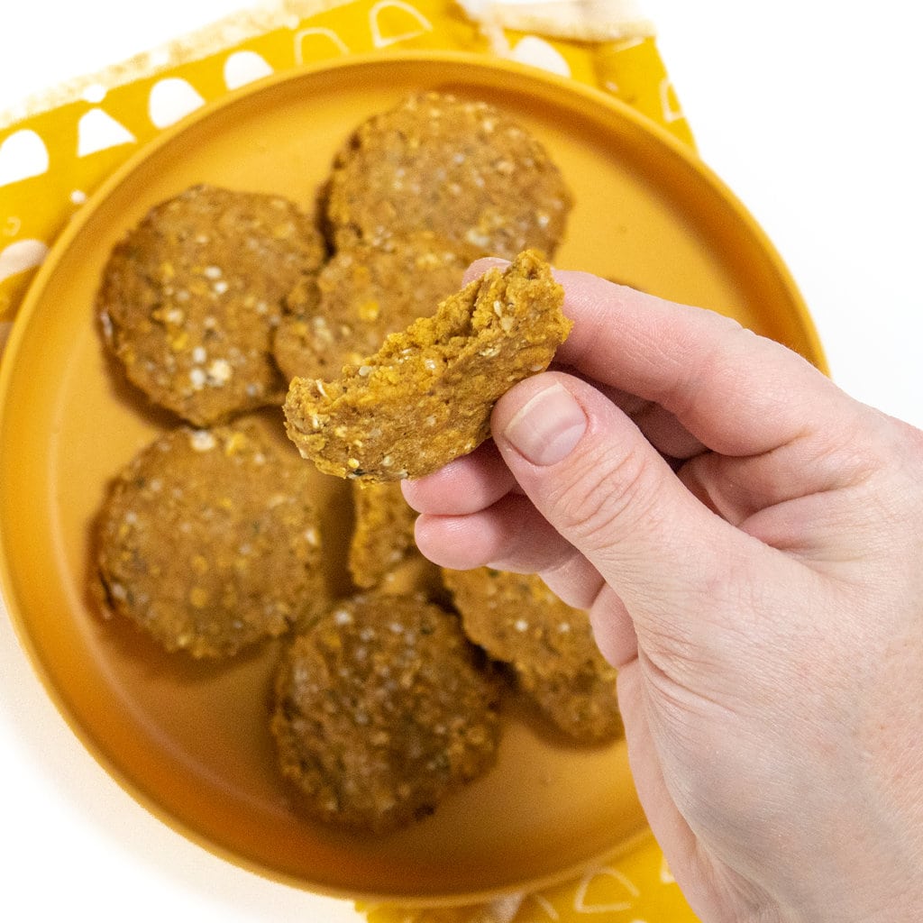A hand holding up a soup, potato baby cookie over a plate of cookies and a mustard color napkin over a white countertop.