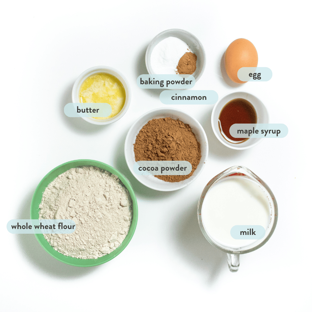 A spread of ingredients for kid, friendly chocolate pancakes, and a white countertop with graphic tags labeling each ingredient.