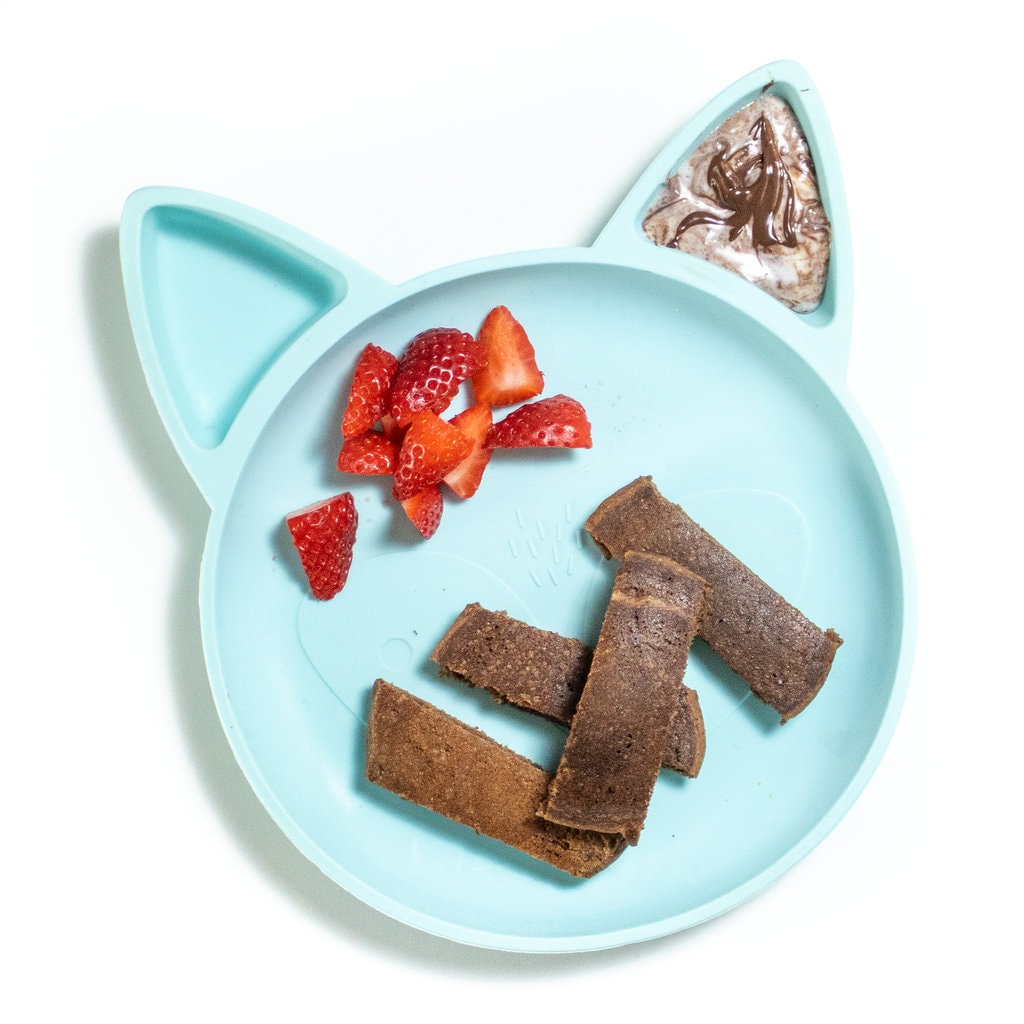 A cat shaped blue toddler plate with a slices of chocolate, pancake, chunks of strawberry, and a yogurt with chocolate drizzle on top.