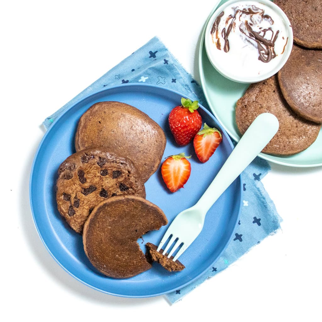 Two blue and teal plates with a blue fork with chocolate pancakes with chocolate chips slice strawberries, yogurt with chocolate sauce, dip, and a white counter with a blue napkin.