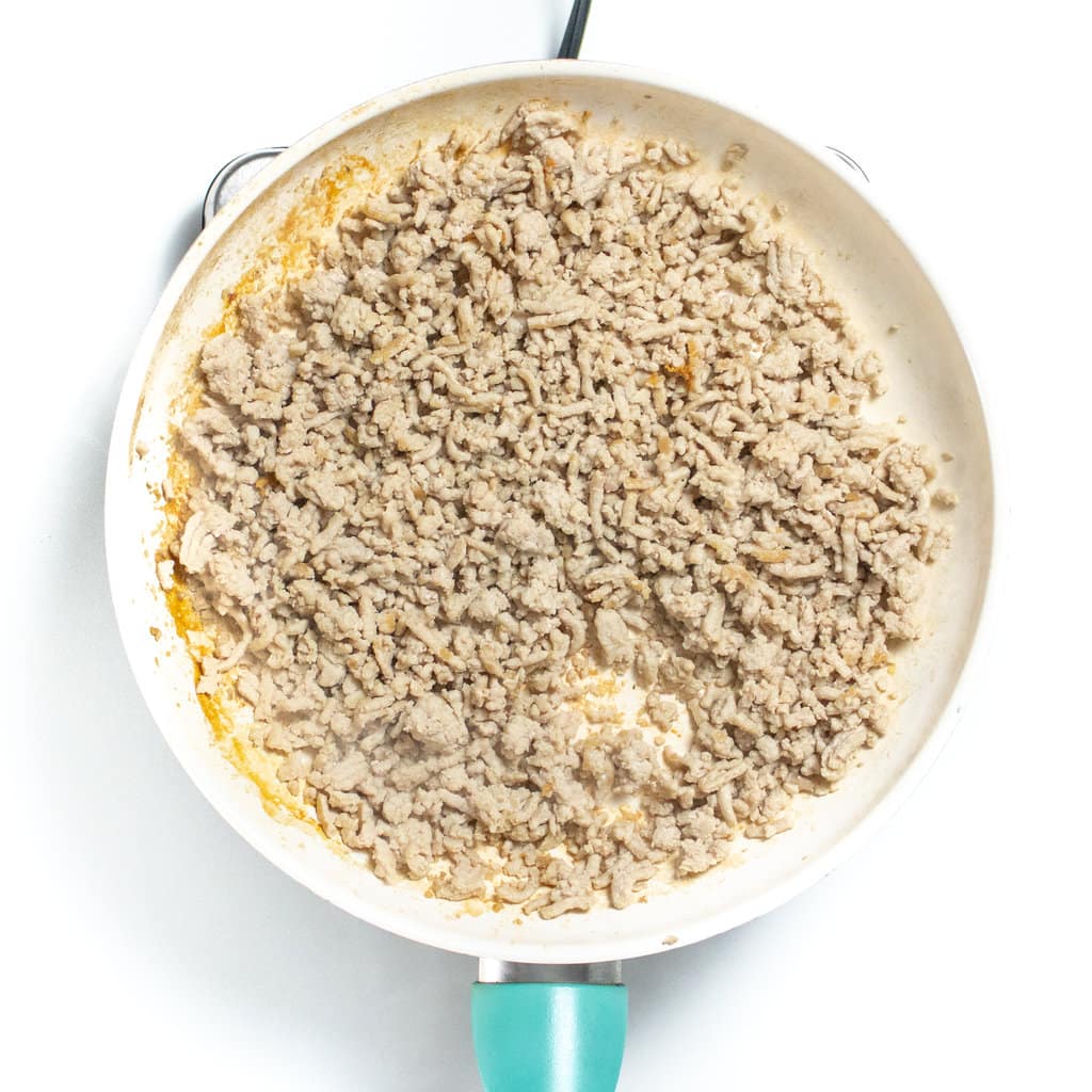 White skillet on a white countertop with ground chicken browned, and still cooking.