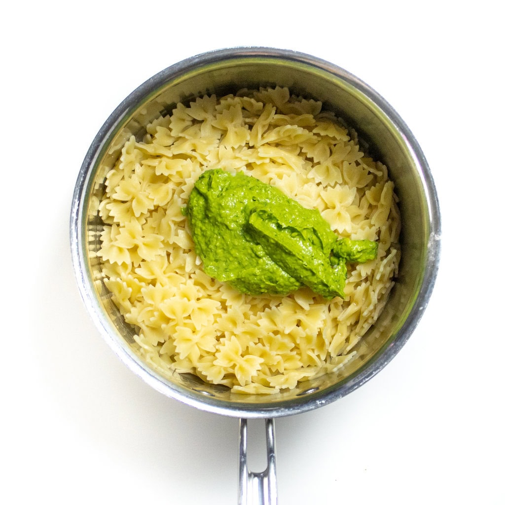 A silver sauce, pan with bowtie, pasta, and a huge spoonful of spinach, avocado pesto on top.