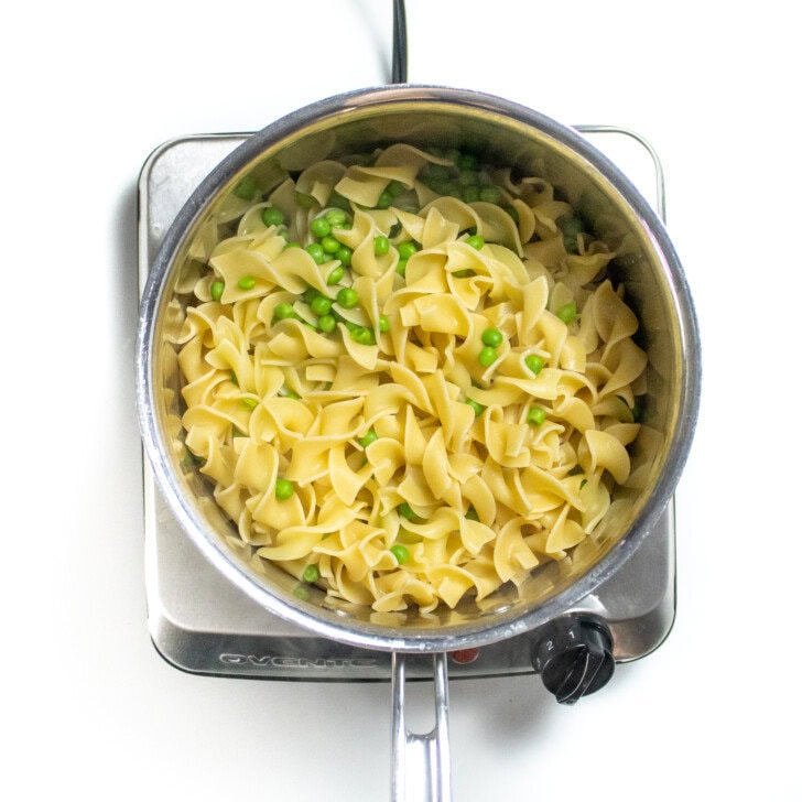 Silver sauce pan on a white countertop with cooked egg noodles and peas.