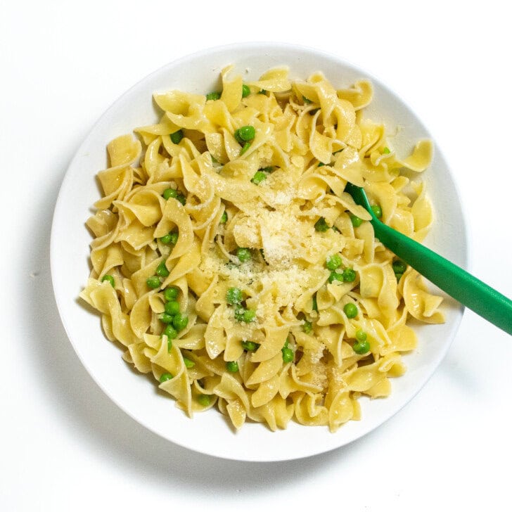 A big white bowl full of butter noodles with peas and Parmesan on a white countertop.