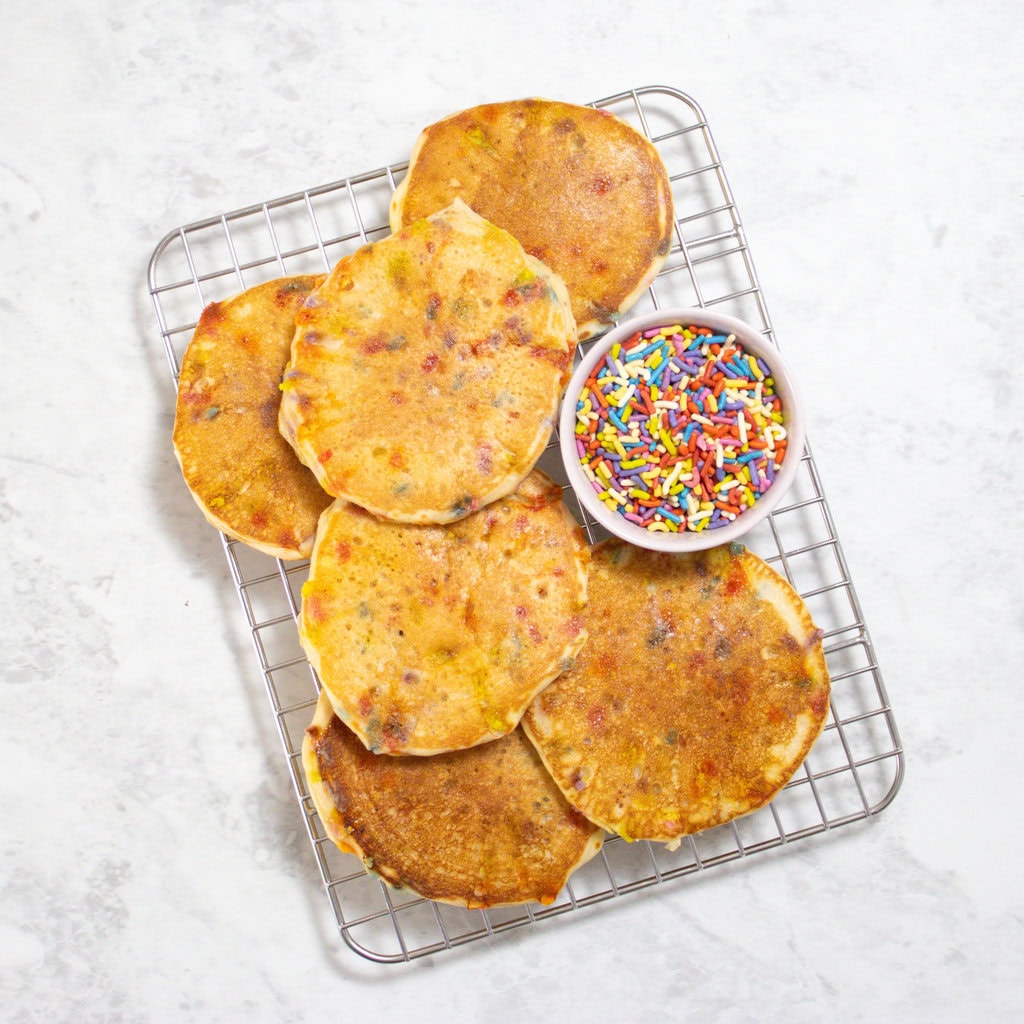 Small white cooling rack with stacks of golden brown funfetti pancakes and a small bowl of rainbow sprinkles.