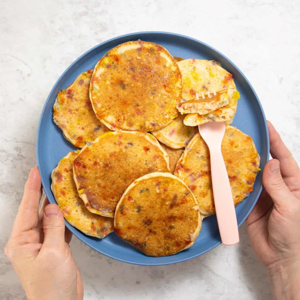 Two hands holding a blue kids plate with stacks of birthday cake pancakes over a white kitchen counter.