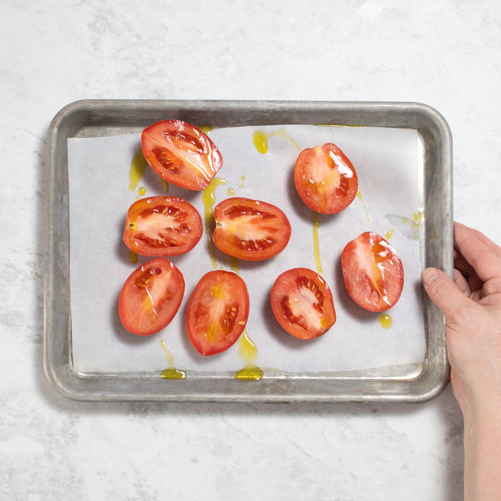 Hand holding a silver tray with cut in half tomatoes with a drizzle of olive oil. Over my marbled white kitchen counter.